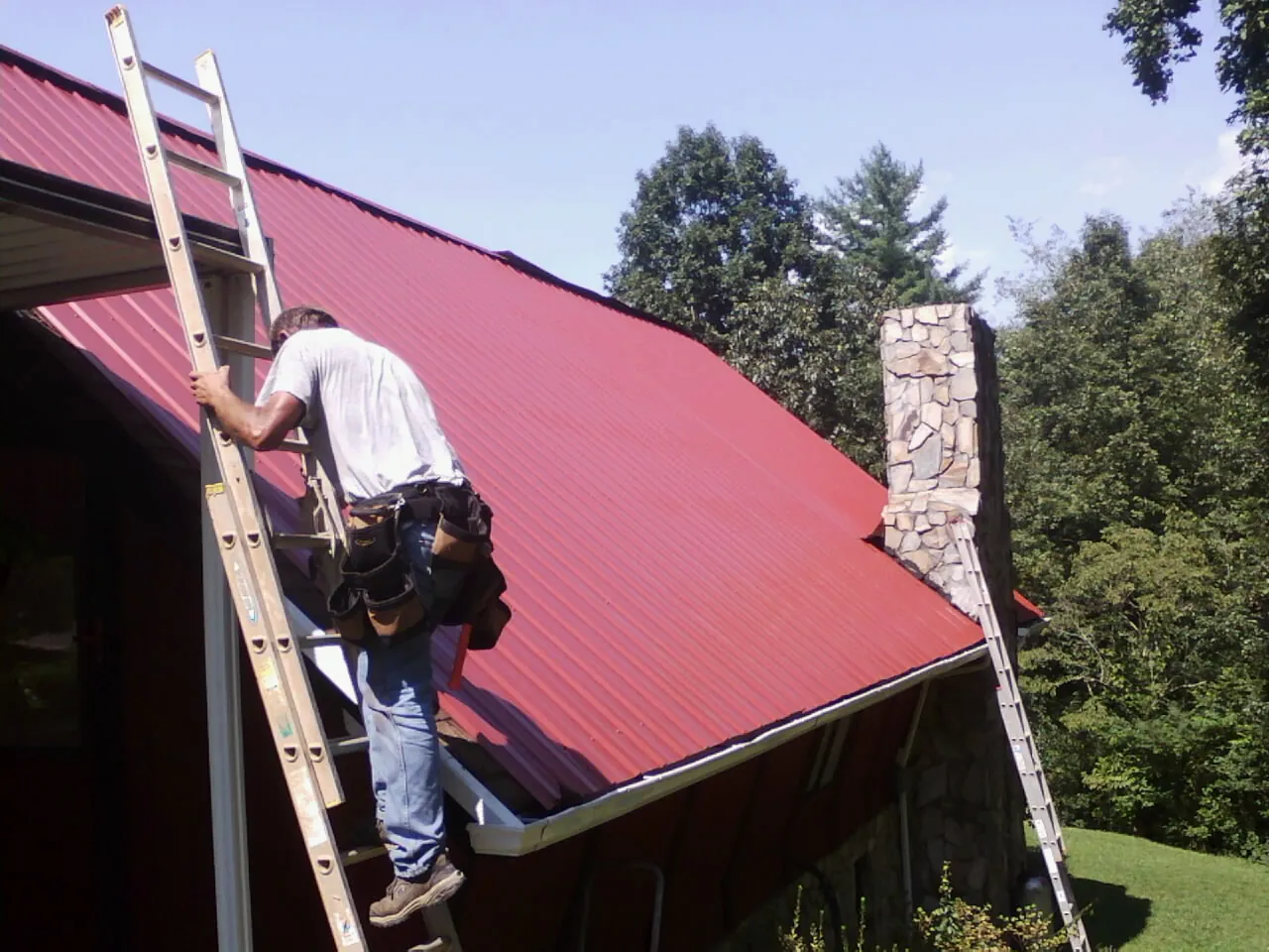 Roofer on a ladder beside a red metal roof and stone chimney — Soffit & Fascia Repair by Roofing Brothers in Perkins