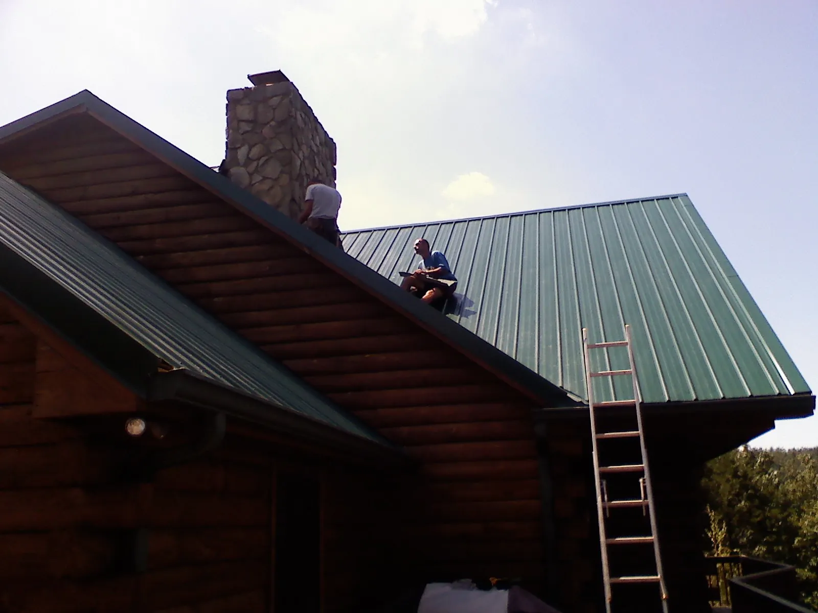 Roofers installing green metal panels beside a stone chimney on a log home in Perkins